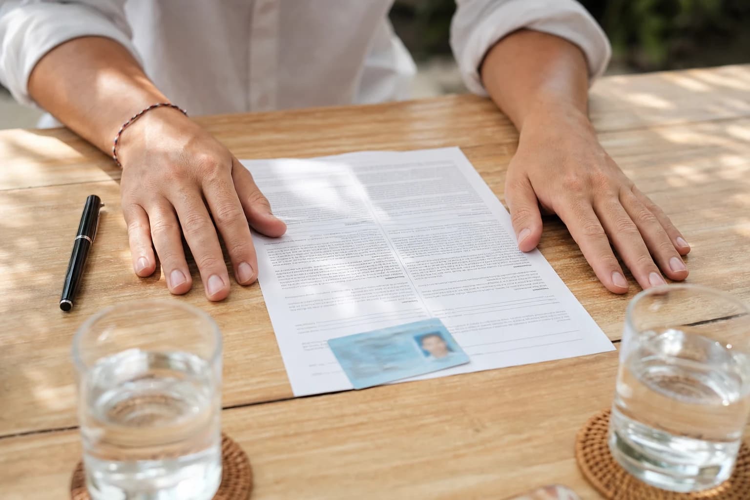 Two hands resting on a pale wooden table above a one-page contract printed in two languages and a blurred Indonesian identity card, with a pen and two glasses of water, natural light from a shaded terrace