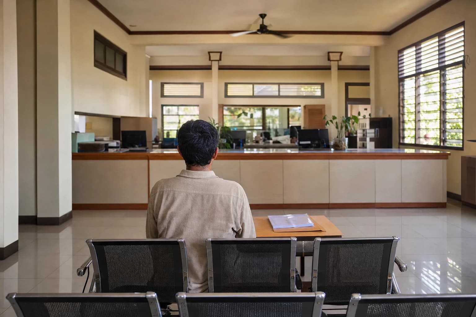 Waiting room of an Indonesian government office in Denpasar, Bali: a person seen from behind, seated facing a formica counter, a cardboard file and a document wallet in front of them, ceiling fan, louvred windows filtering the light, rows of chairs and pale floor tiles, a calm and impersonal atmosphere