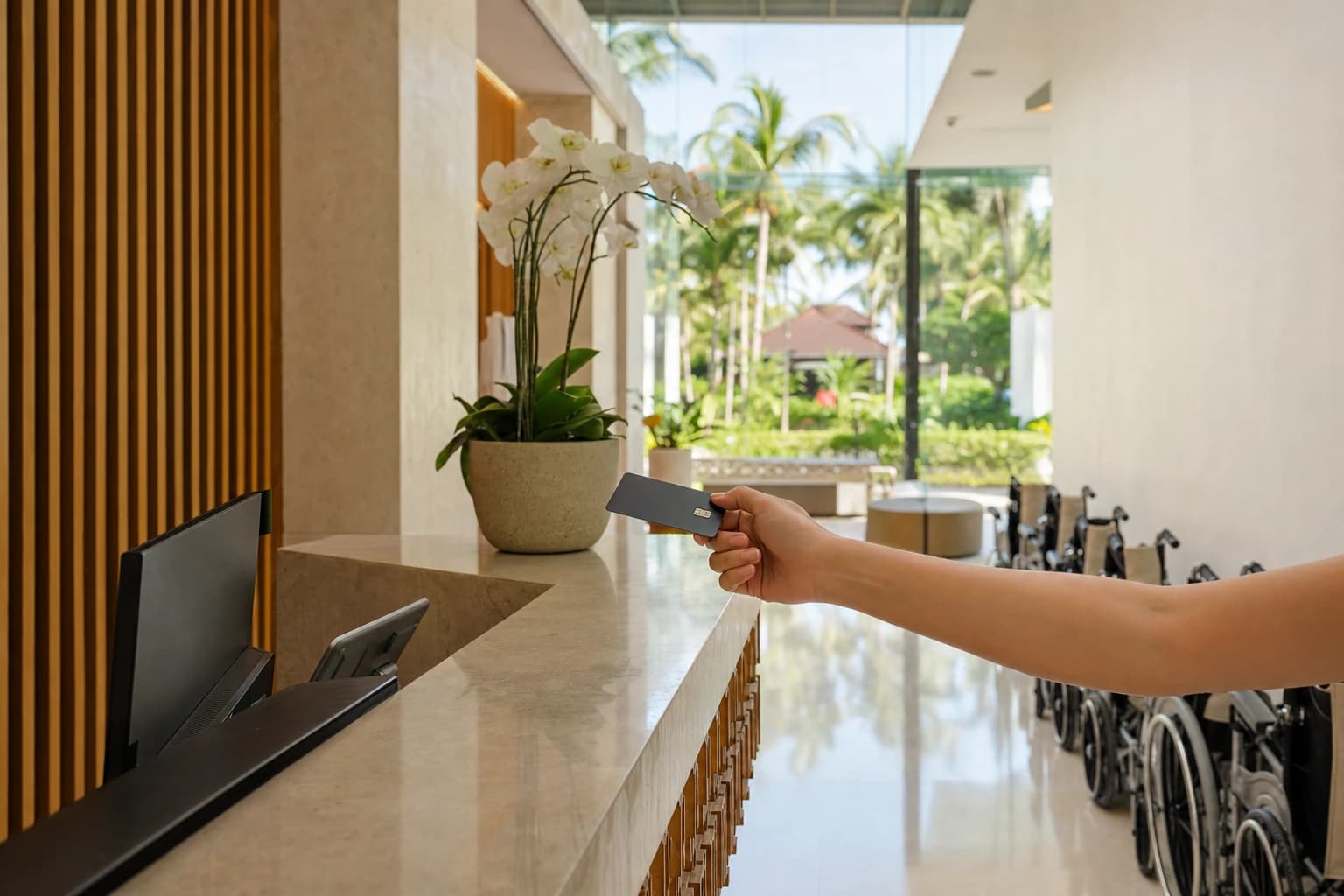 Reception desk of a private hospital in Bali seen from a three-quarter angle, a hand holding out a bank card above the pale stone counter, folded wheelchairs along a white wall, palm trees visible through the glass wall, midday tropical light, no faces in frame, no readable text