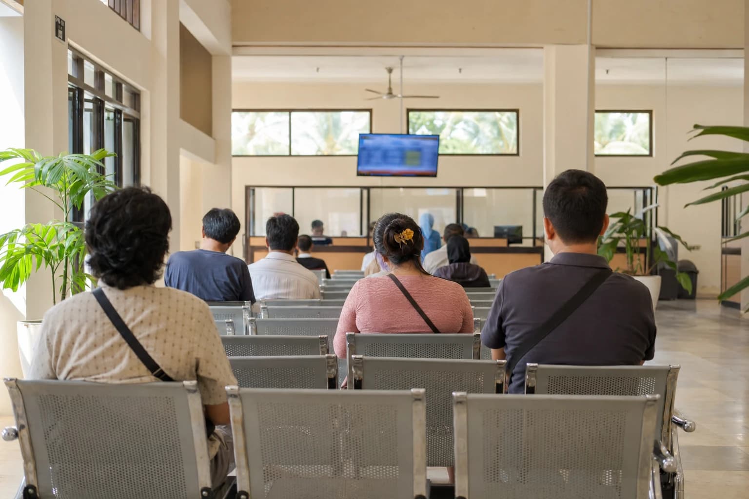 The waiting room of an Indonesian immigration office photographed from behind, rows of metal chairs, a blurred queue-number screen, glazed counters at the far end, ceiling fan, pale tiled floor, potted plants, natural light from tall windows, people seen only from behind