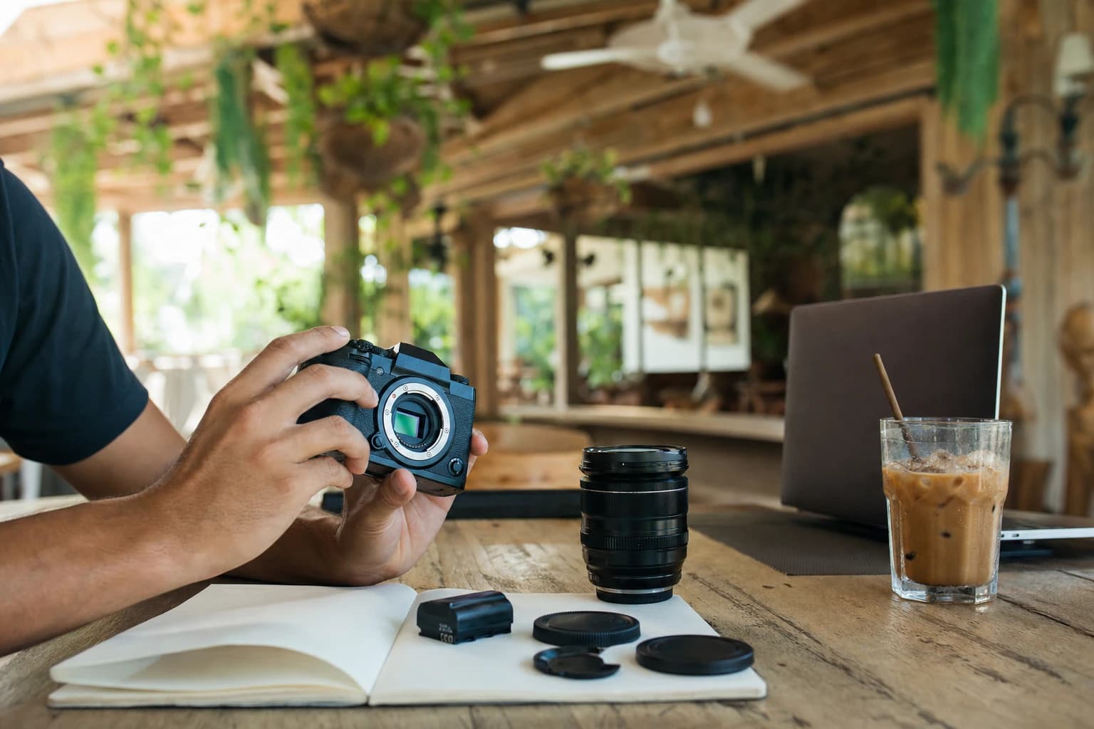 Plan serré sur des mains inspectant un appareil photo hybride noir, objectif détaché posé sur une table de café en bois clair à Canggu, batterie de rechange et capuchons alignés sur un carnet ouvert, ordinateur portable entrouvert à côté d'un verre de café glacé, plantes suspendues et ventilateur de plafond flous en arrière-plan, lumière naturelle de matinée tropicale