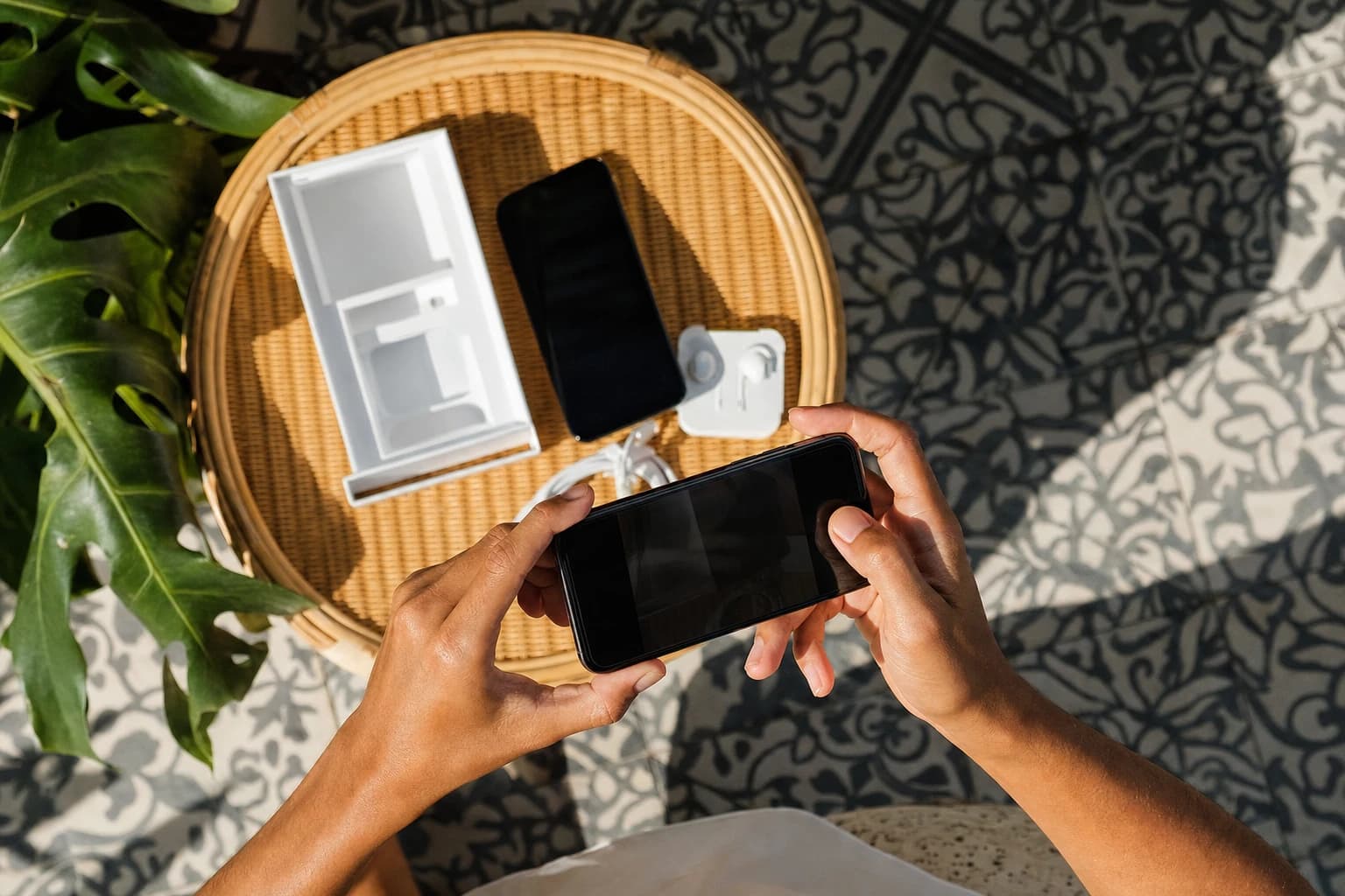 Vue en plongée d'un smartphone photographié par un second téléphone tenu à deux mains au-dessus d'un plateau en rotin clair, boîte d'origine ouverte, câble enroulé et écouteurs disposés au carré sur un sol en carreaux de ciment balinais, ombres nettes de fin de matinée, feuilles de monstera entrant dans un coin du cadre