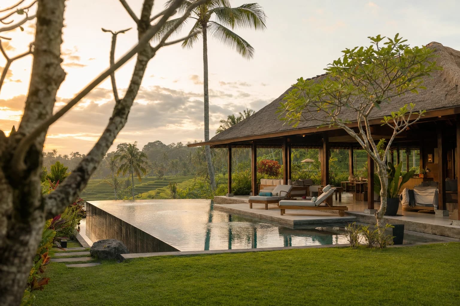 A contemporary Balinese villa with a thatched roof seen from the garden, infinity pool edged in grey volcanic stone, palms and frangipani trees, terraced rice fields blurred in the background, late-afternoon sky
