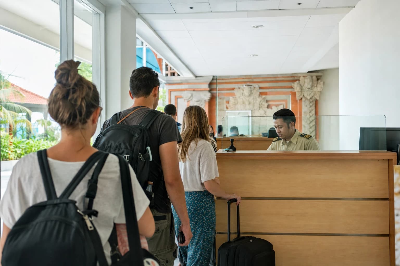 Indonesian customs counter in an airport terminal in Bali, pale wood desk with a screen and a stamp sitting on the worktop, a short queue of travellers seen from behind with suitcases, cold fluorescent light, white walls