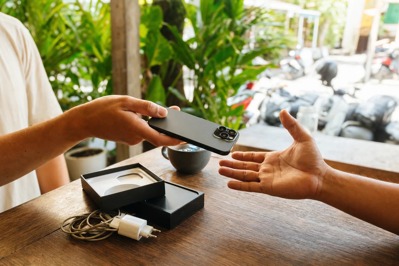 Two hands over a wooden table in an open-air cafe in Canggu, one passing a second-hand smartphone to the other, original box and charger beside them, tropical plants and blurred scooters in the background, natural mid-morning light