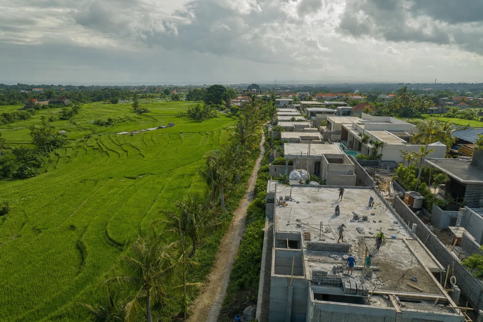 Oblique aerial view over Canggu showing the sharp boundary between bright-green terraced rice fields and a row of new flat-roofed villas with turquoise pools, a dirt subak path, coconut palms, a few tiny construction workers seen from above on a concrete slab, heavy tropical sky, no text or signs