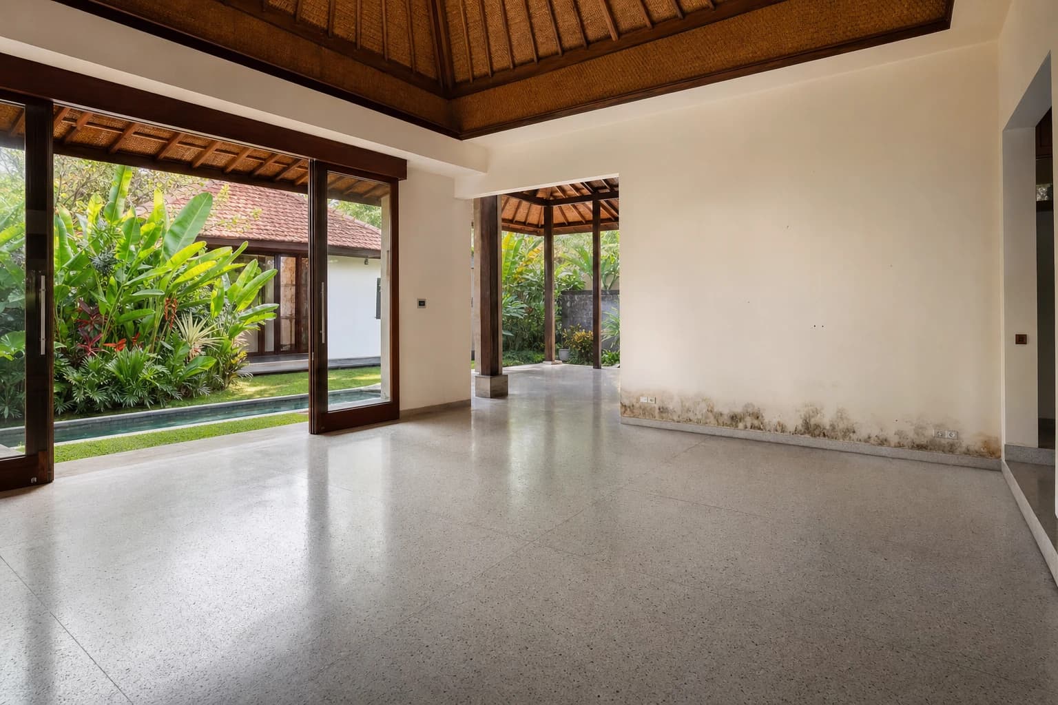 The living room of an empty, unfurnished Balinese villa, grey terrazzo floor, large sliding glass doors open onto a tropical garden, damp marks along the bottom of a white wall, natural morning light