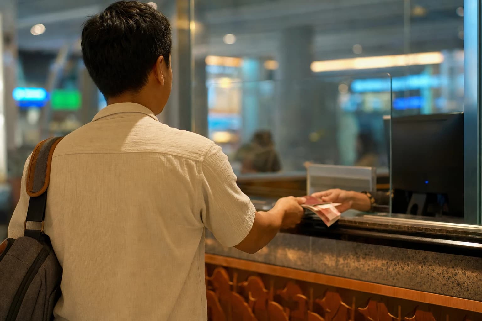Immigration counter in the departures hall of Ngurah Rai airport in Denpasar, seen from a rear three-quarter angle: a traveller from behind, bag over one shoulder, handing a passport and a wad of 100,000-rupiah notes through a glass counter window, blurred signage in the background, neon lighting, no faces visible