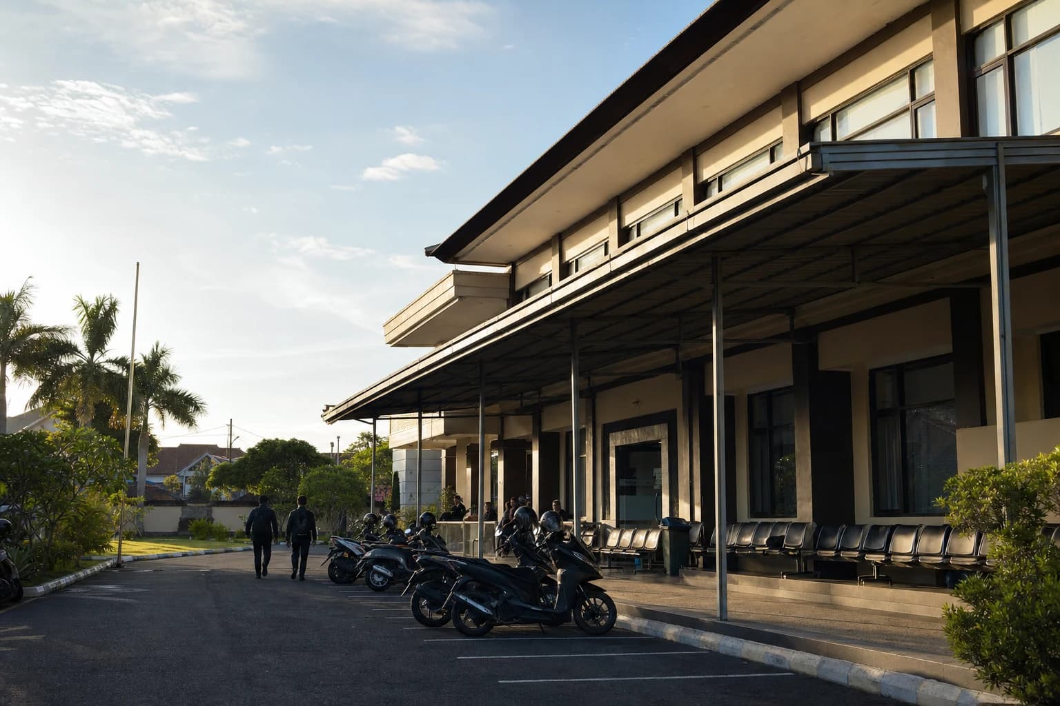 Indonesian police administrative building in Denpasar in the early morning, beige facade and metal canopy, a row of empty plastic chairs beneath it, distant figures seen from behind with no identifiable faces, scooters parked at an angle in front of the entrance, faintly hazy sky