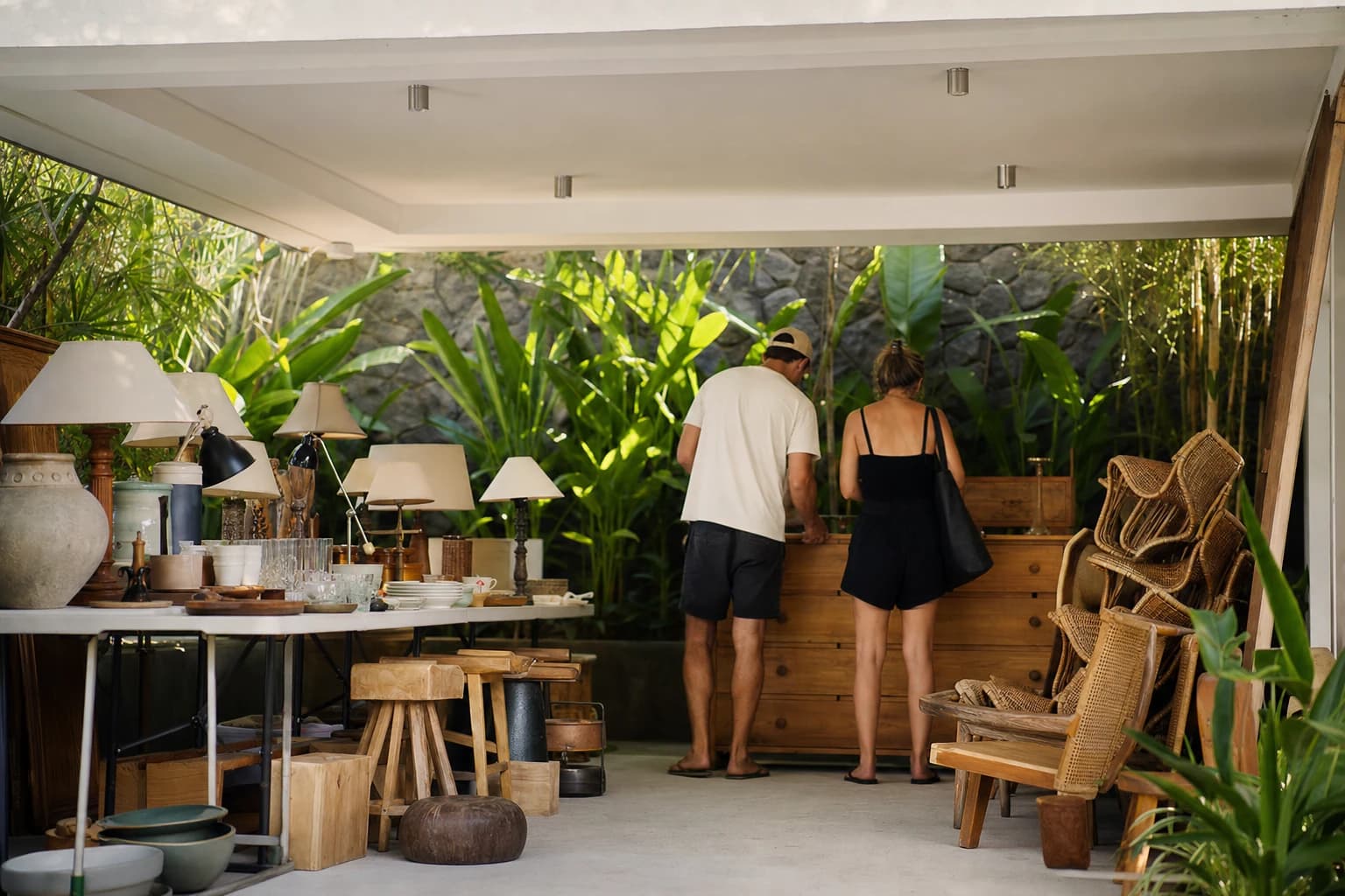 Un vide-maison improvisé dans le carport d'une villa de Canggu : tables pliantes couvertes de lampes, vaisselle et petit mobilier en teck, pile de chaises en rotin sur le côté, deux acheteurs vus de dos qui examinent une commode, végétation tropicale et mur en pierre volcanique en arrière-plan, lumière de fin d'après-midi