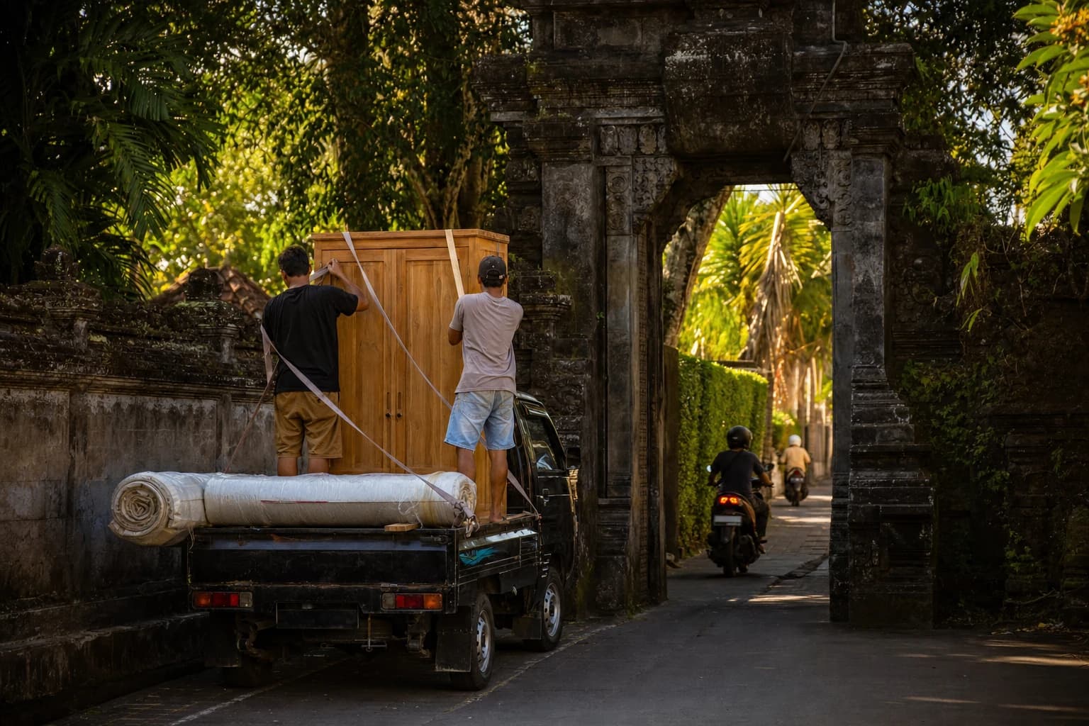 Un pickup à plateau ouvert stationné devant un portail balinais en pierre sculptée, deux personnes vues de dos en train de sangler une armoire en teck et un matelas roulé sur le plateau, scooters passant dans la ruelle étroite bordée de murs végétalisés, lumière chaude de fin de journée