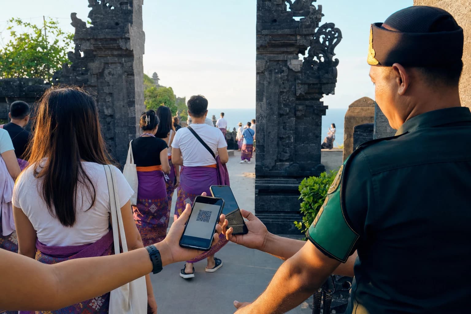 Entrance to a Balinese temple on a cliff facing the ocean, queue of visitors in sarongs seen from behind, uniformed officer scanning the QR code on a smartphone held by a visitor of whom only the hand is visible, carved volcanic stone gates, late-afternoon sky, no faces