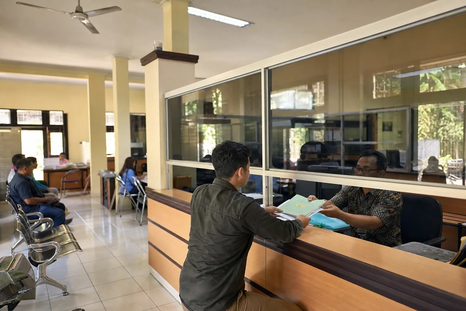 An Indonesian land office counter seen from three-quarters behind, a visitor with their back turned handing a file and a green-covered certificate to a clerk behind glass, blurred signage, rows of waiting chairs, strip lights and a ceiling fan, tropical administrative atmosphere